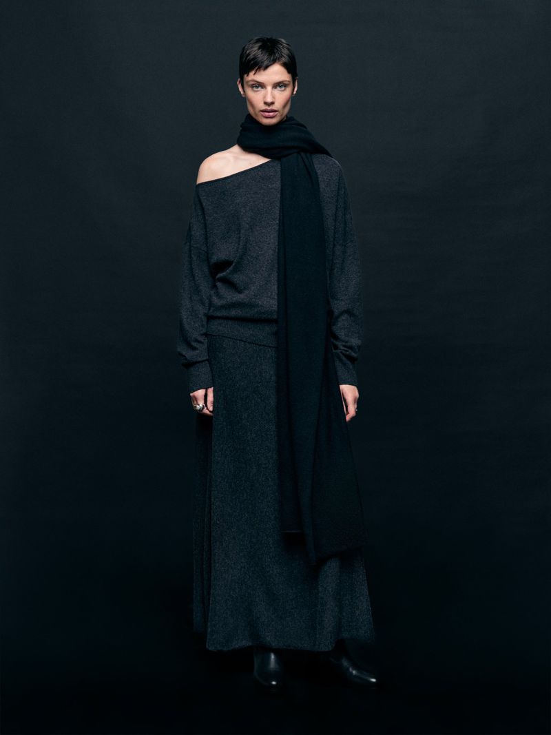 Fashionable adult woman in a studio portrait wearing a dark sweater, scarf, and skirt posing against a dark backdrop with eye-level camera angle.