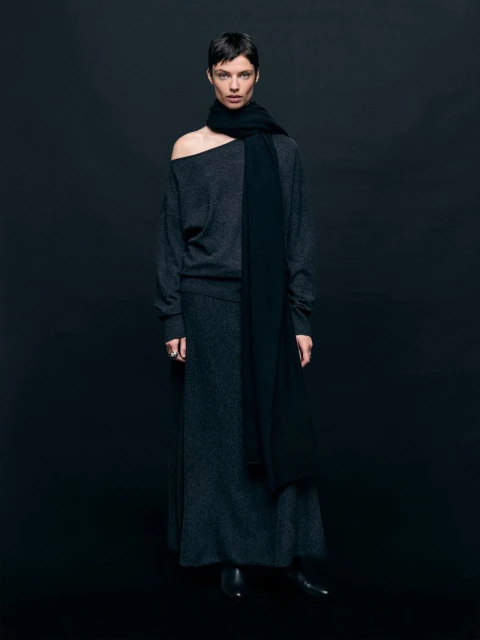 Fashionable adult woman in a studio portrait wearing a dark sweater, scarf, and skirt posing against a dark backdrop with eye-level camera angle.