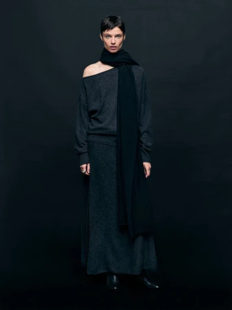 Fashionable adult woman in a studio portrait wearing a dark sweater, scarf, and skirt posing against a dark backdrop with eye-level camera angle.