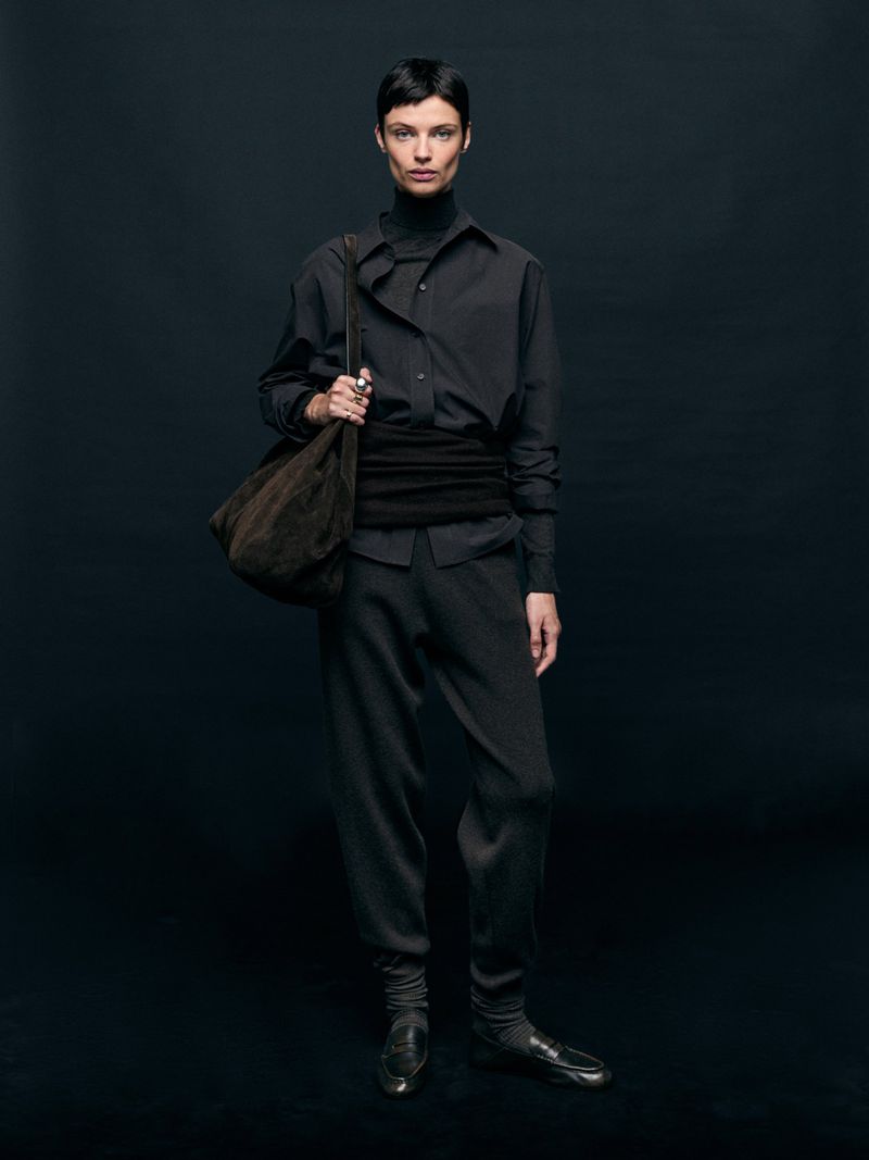 Fashionable adult female in all-black outfit posing in a photo studio with a soft brown bag; full-length portrait on a dark background.