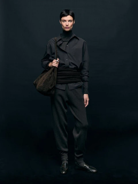Fashionable adult female in all-black outfit posing in a photo studio with a soft brown bag; full-length portrait on a dark background.