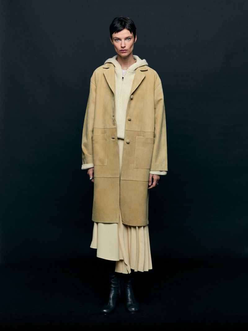 Adult woman wearing a tan suede coat, cream-colored hoodie and skirt, and black leather boots, posing in front of a dark background.