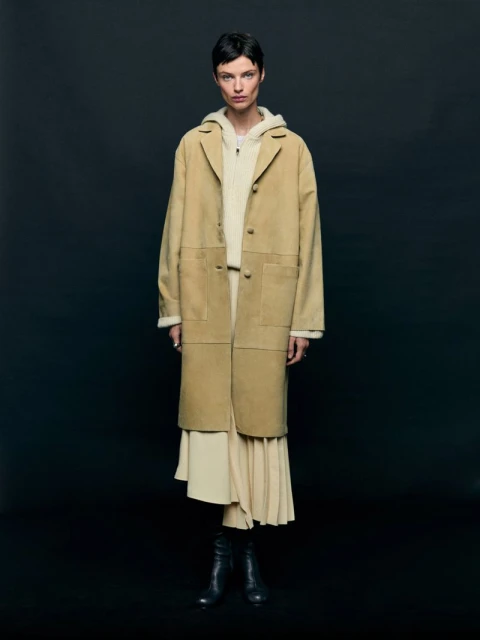 Adult woman wearing a tan suede coat, cream-colored hoodie and skirt, and black leather boots, posing in front of a dark background.