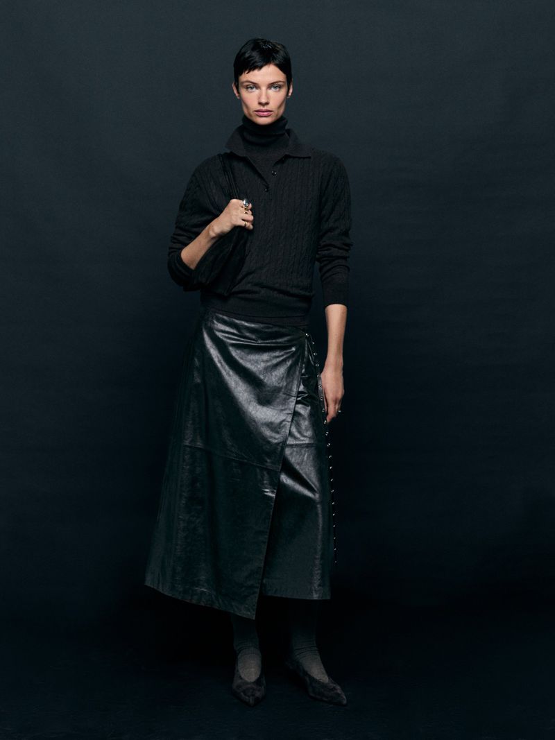 A woman wearing a black sweater and a leather wrap skirt stands in a studio with a black background, carrying a matching black bag.