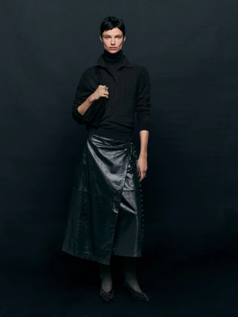 A woman wearing a black sweater and a leather wrap skirt stands in a studio with a black background, carrying a matching black bag.