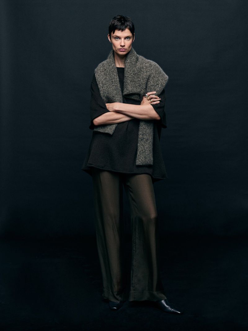 Full-length portrait of a woman in sheer pants and a draped scarf against a dark background in a fashion-forward studio shot.