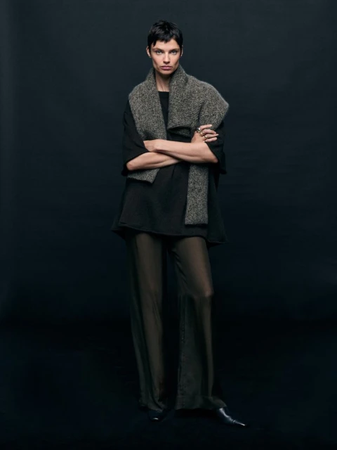 Full-length portrait of a woman in sheer pants and a draped scarf against a dark background in a fashion-forward studio shot.