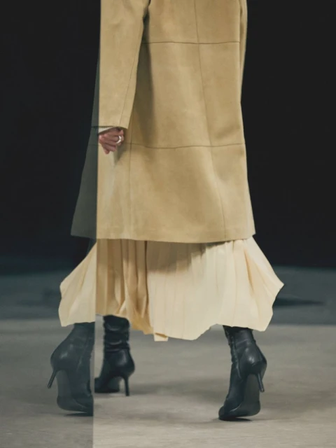 Fashionable model showcases a beige coat, pleated skirt, and black heeled boots against a dark background in a studio fashion shoot.