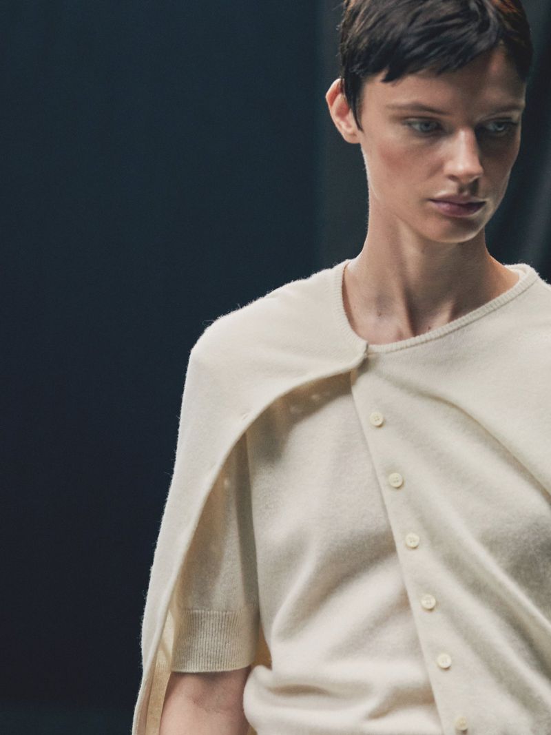 Woman with short hair poses indoors wearing a cream-colored cardigan and shirt. Focus on fashion and the neutral color palette.