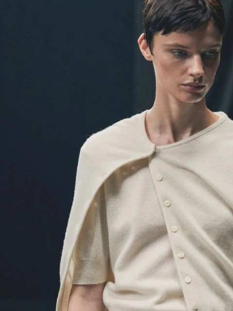 Woman with short hair poses indoors wearing a cream-colored cardigan and shirt. Focus on fashion and the neutral color palette.