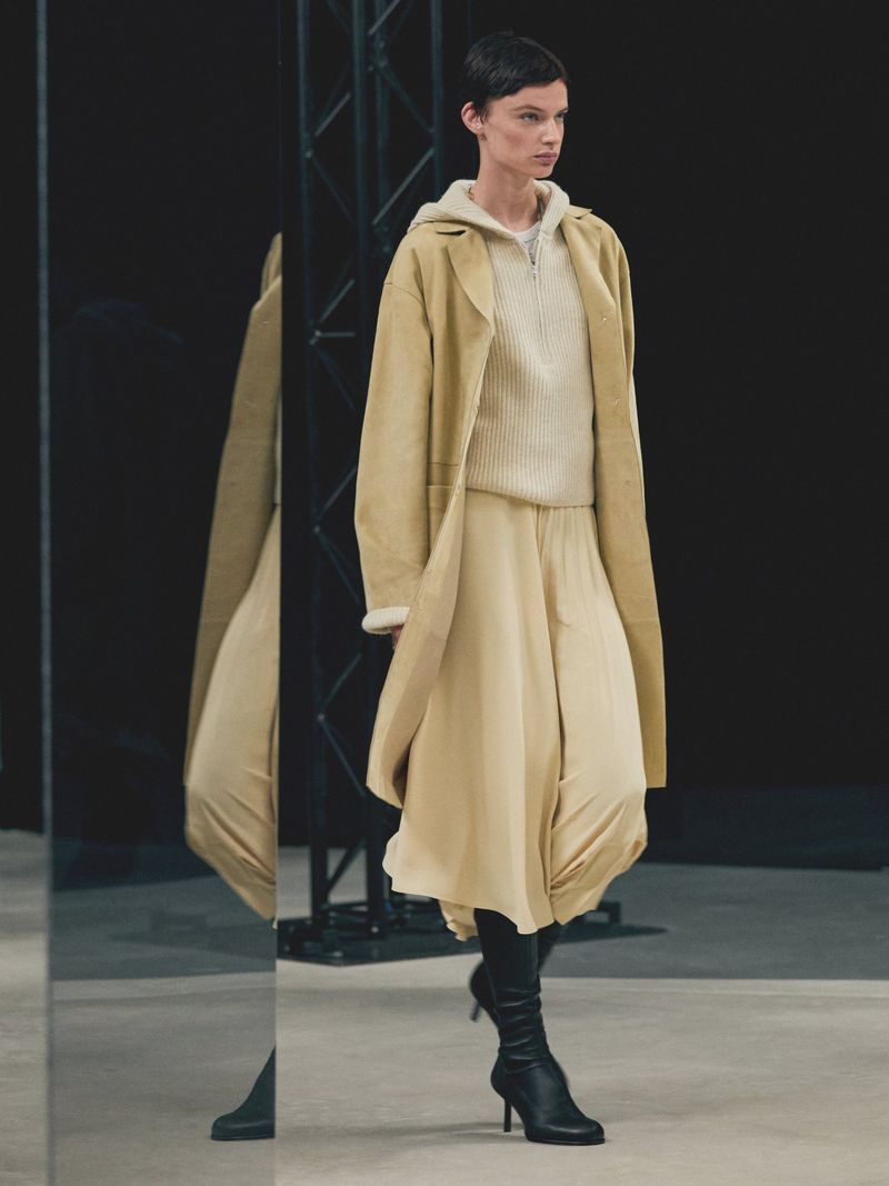 A woman models a beige outfit with a long coat, sweater, and skirt, paired with black heeled boots on a light surface with a black background.