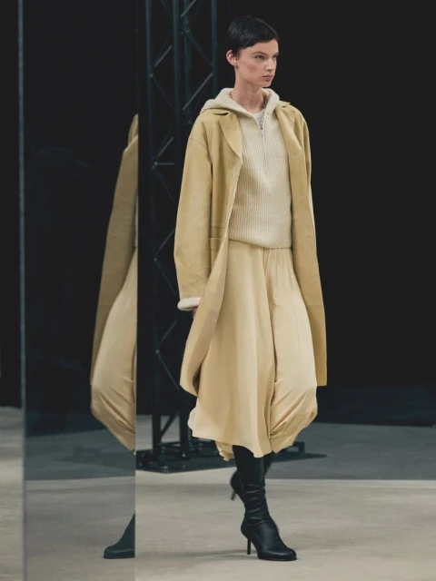 A woman models a beige outfit with a long coat, sweater, and skirt, paired with black heeled boots on a light surface with a black background.