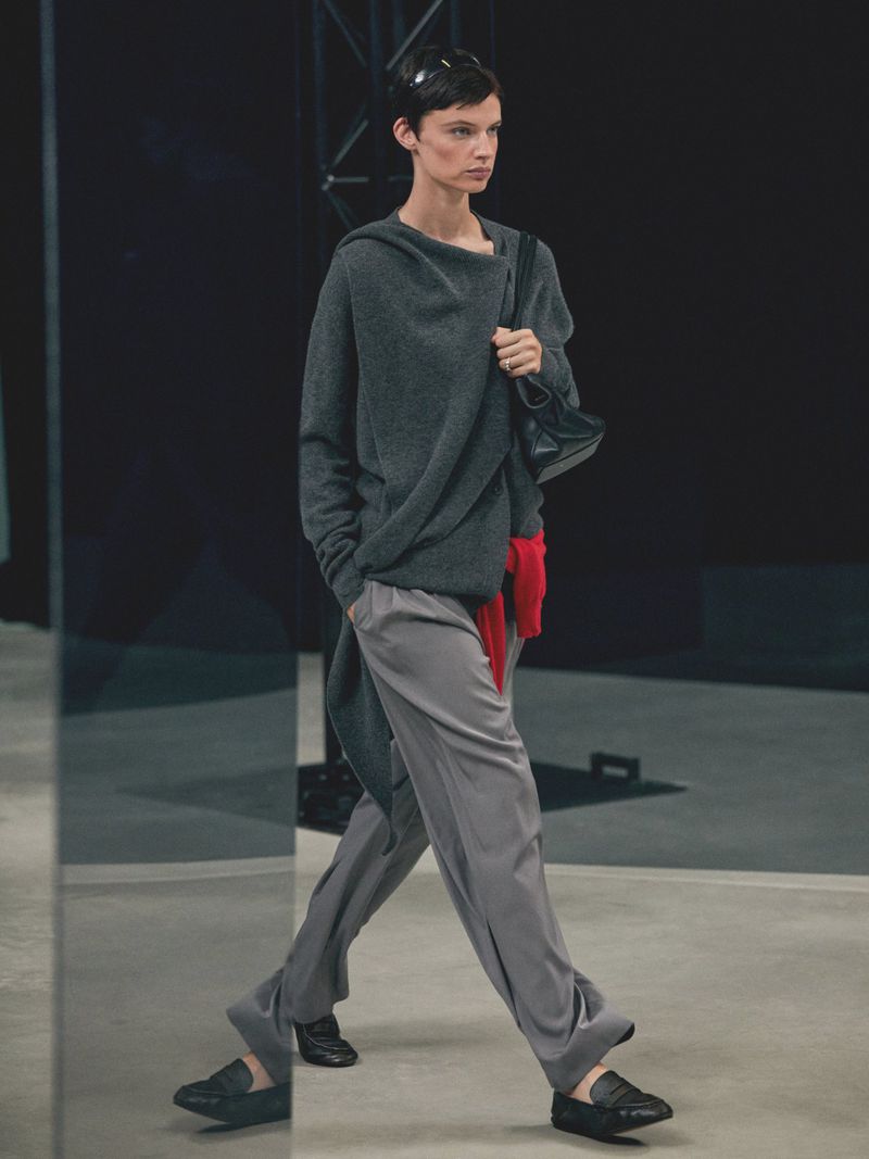 An adult female model walks the runway wearing a grey top, grey pants, and black shoes, carrying a black handbag, indoors.
