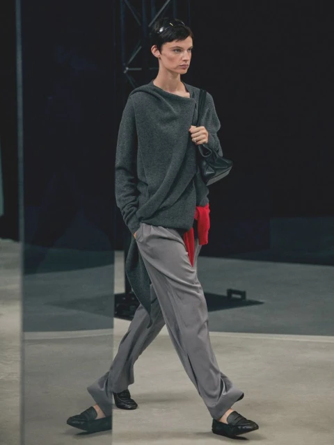 An adult female model walks the runway wearing a grey top, grey pants, and black shoes, carrying a black handbag, indoors.