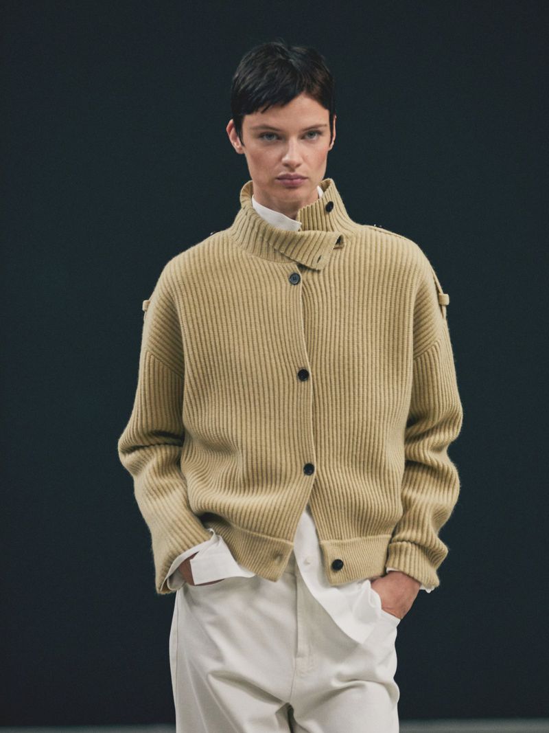 Model wearing a ribbed beige cardigan with a high buttoned collar over a white shirt against a dark background
