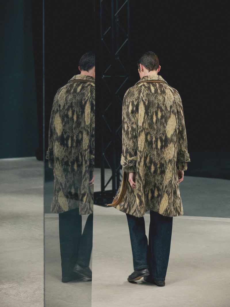 Back view of an adult male in a snakeskin-patterned coat and jeans, standing near a mirror in a photo studio against a black backdrop.