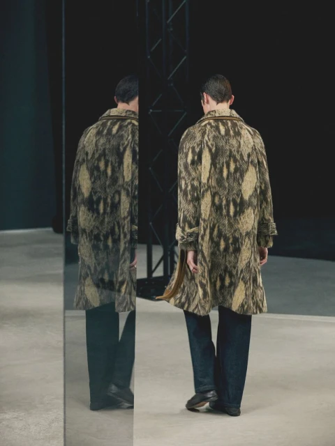 Back view of an adult male in a snakeskin-patterned coat and jeans, standing near a mirror in a photo studio against a black backdrop.