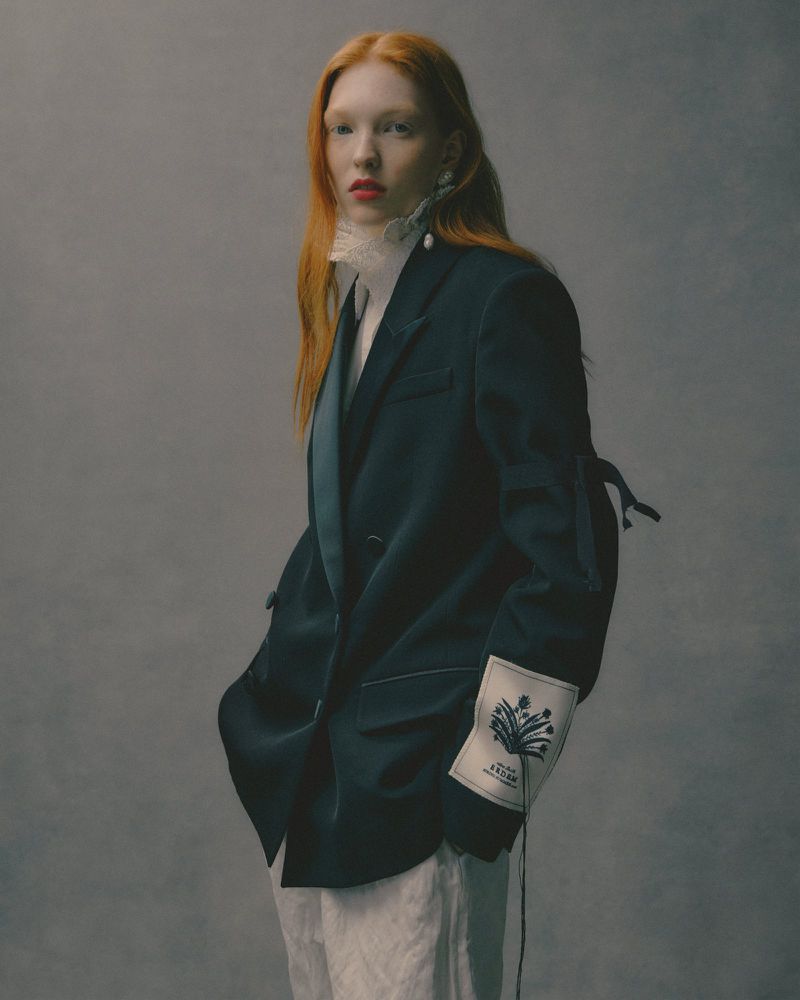 Portrait of a red-haired woman in an oversized blazer and ruffled collar, posing against a muted grey backdrop in a fashion editorial style.