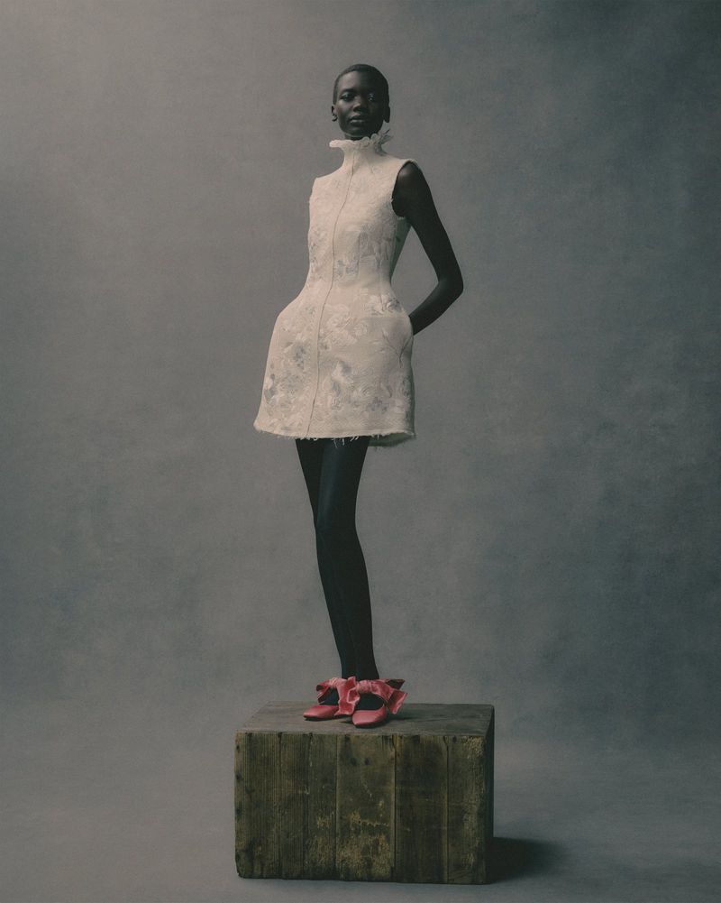 Fashion shot of Black woman in a short white dress with a high collar and pink heels, standing on a rustic wooden box in studio.