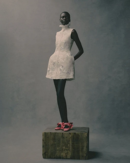 Fashion shot of Black woman in a short white dress with a high collar and pink heels, standing on a rustic wooden box in studio.