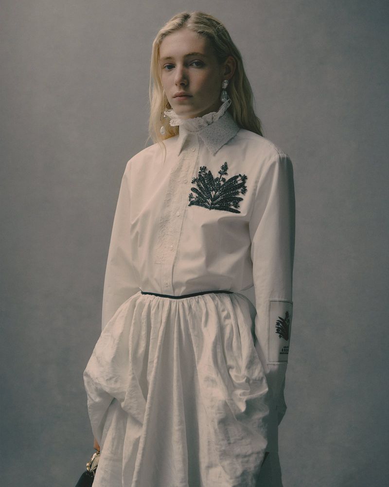 Portrait of a fair-skinned woman with blonde hair wearing a white ruffled high-neck button-down shirt and skirt with embellishments in a studio setting.