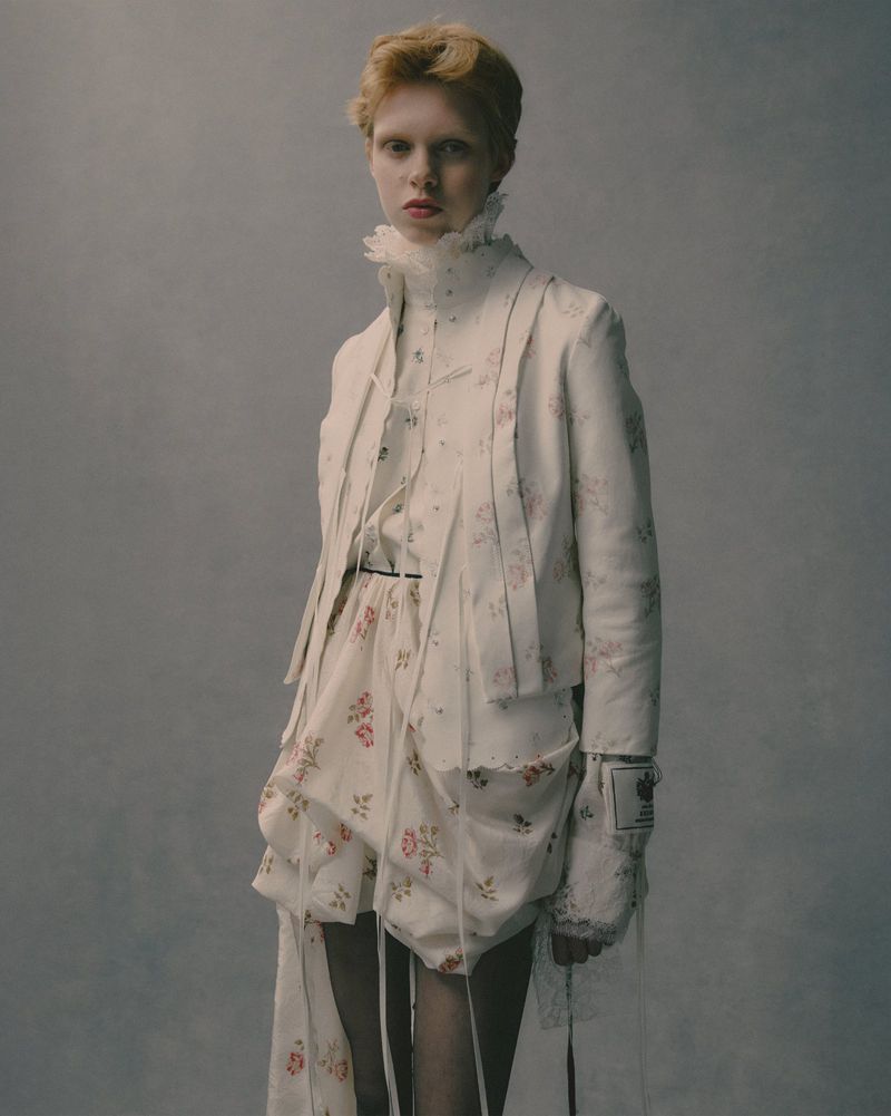Young woman in a white floral jacket and shorts poses in a studio with neutral background. Fashion editorial portrait.