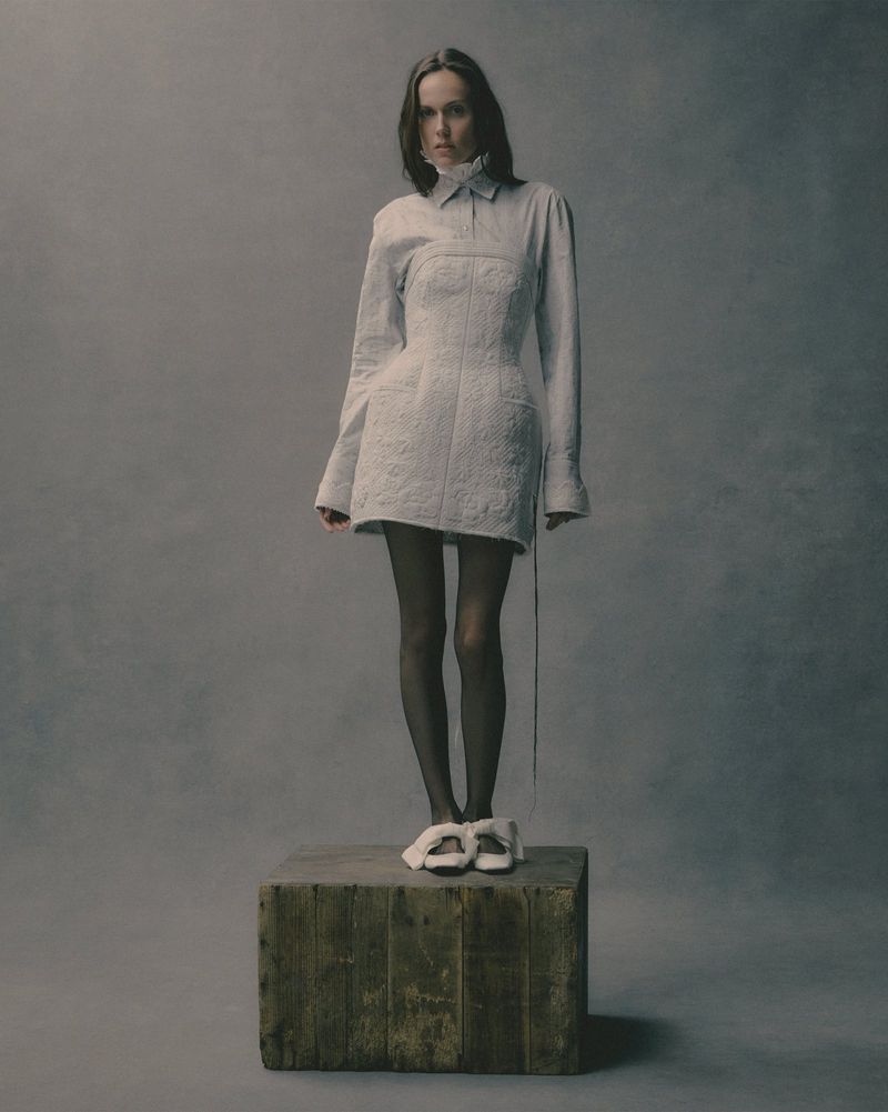 Fashion shot of a woman in a white dress standing on a wooden box in a studio, showcasing an editorial style and neutral color palette.