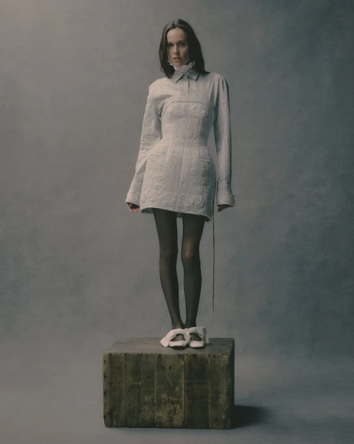 Fashion shot of a woman in a white dress standing on a wooden box in a studio, showcasing an editorial style and neutral color palette.