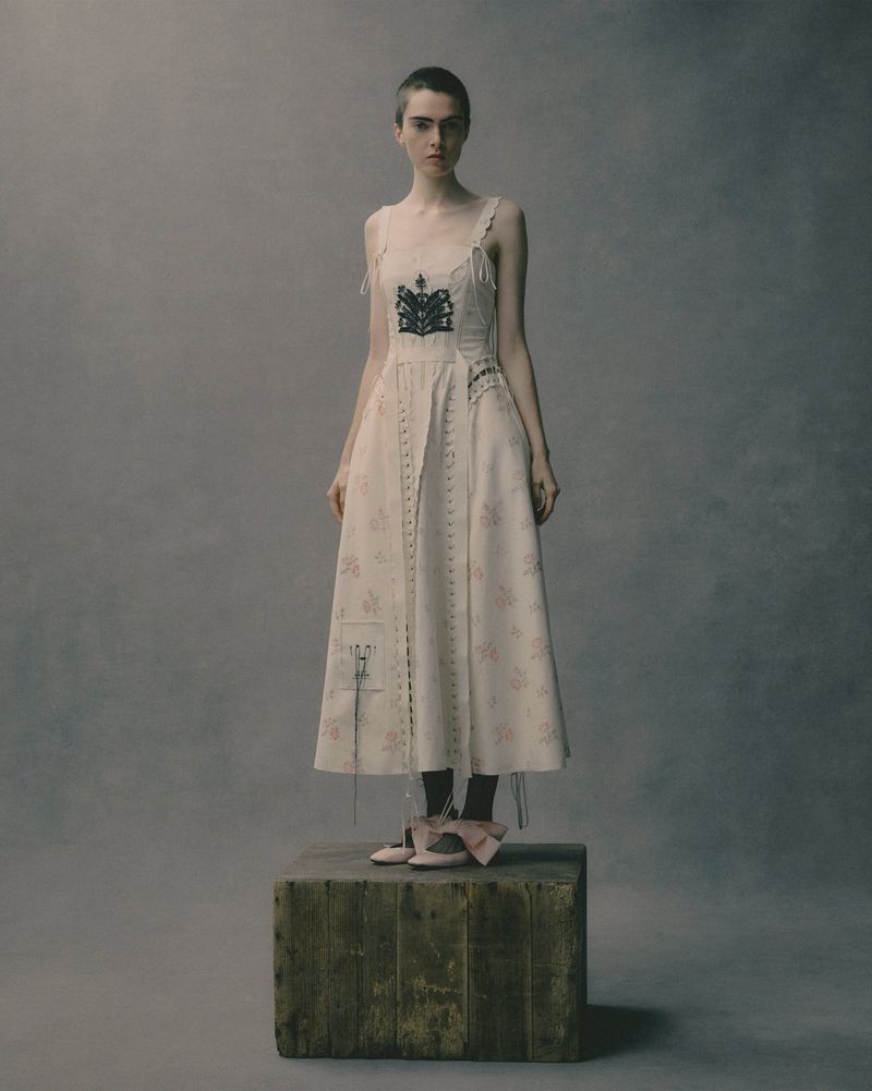 A person in a vintage-inspired dress poses on a wooden block against a mottled gray backdrop, creating a timeless and artistic image.