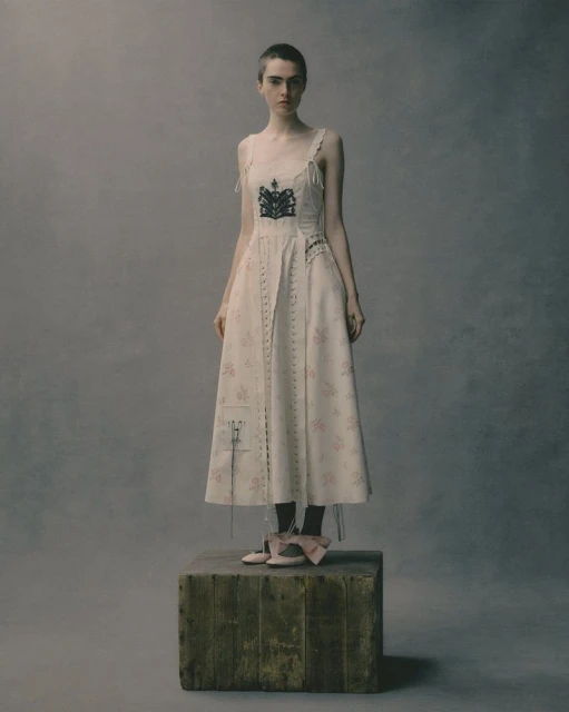 A person in a vintage-inspired dress poses on a wooden block against a mottled gray backdrop, creating a timeless and artistic image.