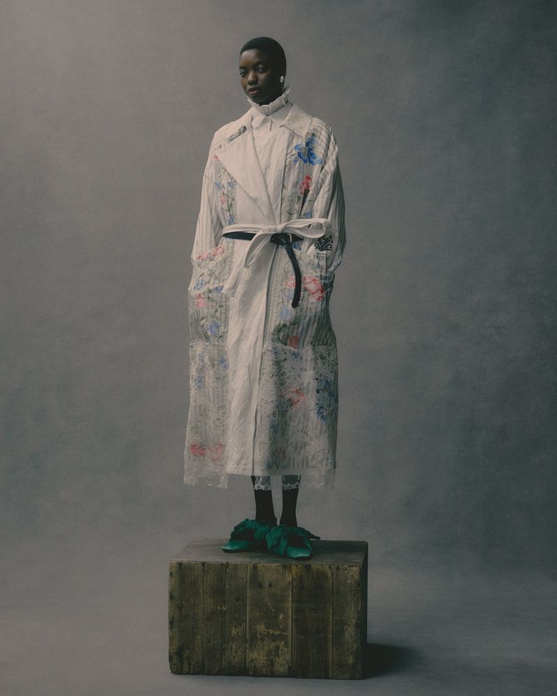 Fashion shot of a Black woman in a floral trench coat standing on a wooden block in a studio, modelling stylish, high fashion.