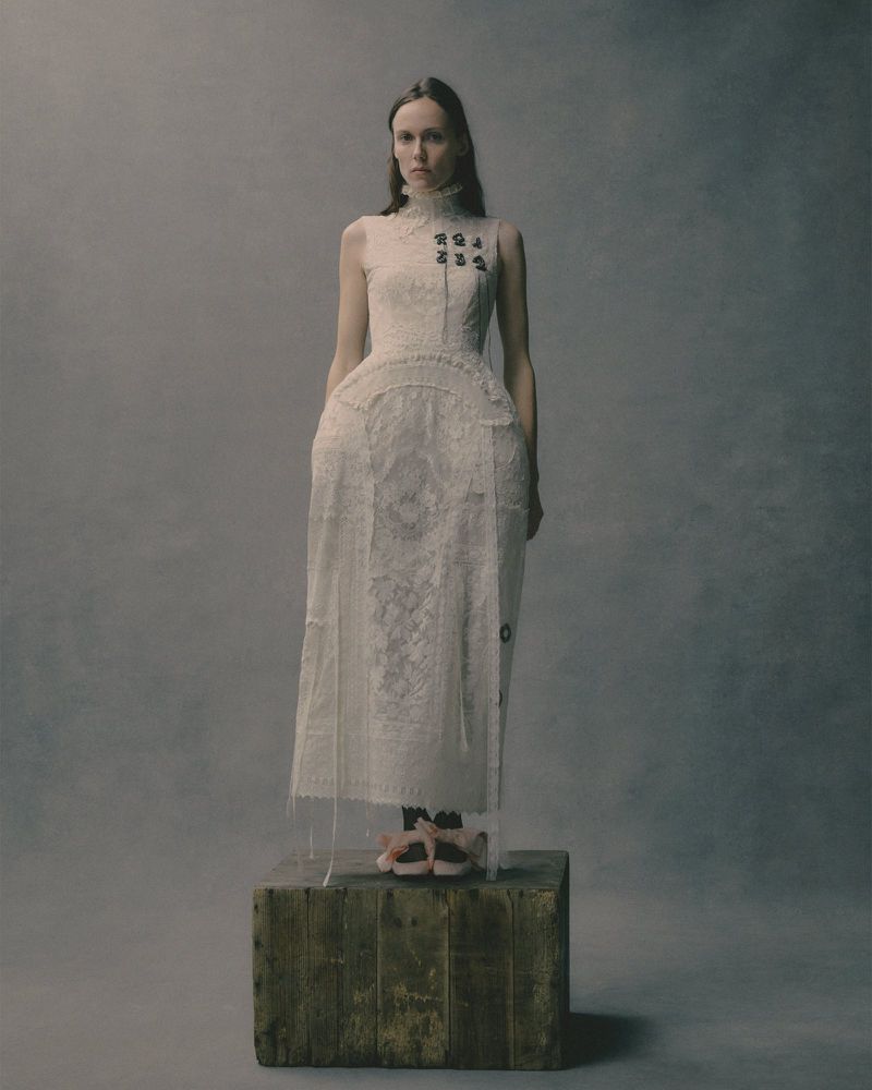 A slender woman posing in a lace dress on a wooden platform, set against a neutral backdrop in a fashion editorial shot.