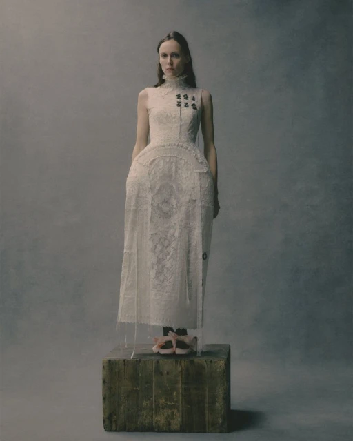 A slender woman posing in a lace dress on a wooden platform, set against a neutral backdrop in a fashion editorial shot.