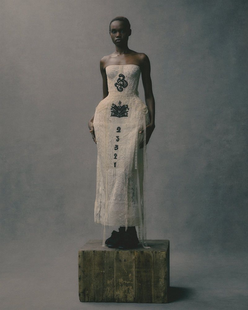 An African American fashion model in a white dress stands on a wooden crate, photographed in a studio with a grey background.