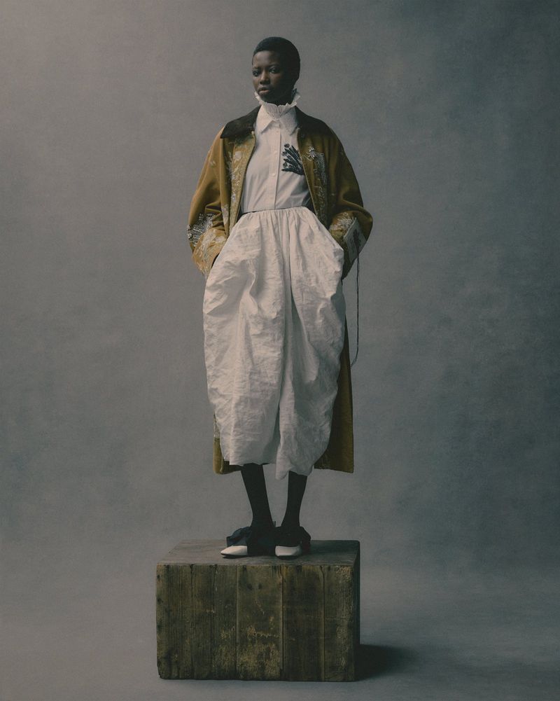 Full-length portrait of a Black woman in a beige coat, white shirt and pants, and dark shoes, posing on a wooden box against a grey background.