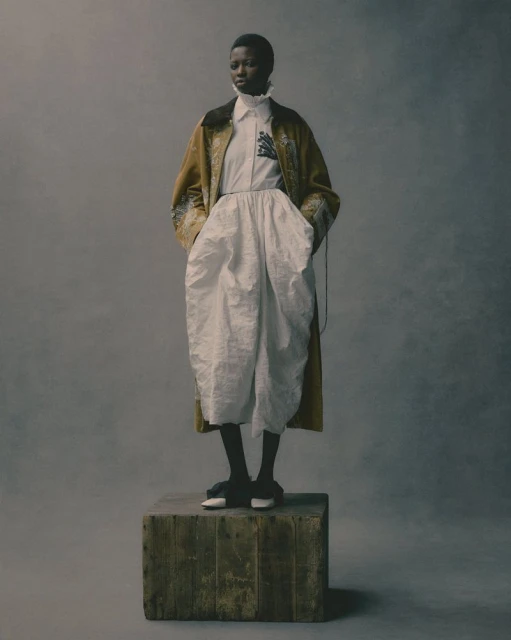 Full-length portrait of a Black woman in a beige coat, white shirt and pants, and dark shoes, posing on a wooden box against a grey background.