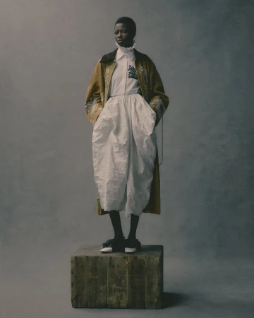 Full-length portrait of a Black woman in a beige coat, white shirt and pants, and dark shoes, posing on a wooden box against a grey background.