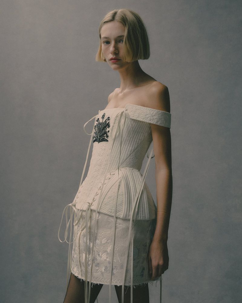 Fashion shot of a young woman with blonde bob wearing a white, off-the-shoulder corset dress with black tights, posing in a studio.