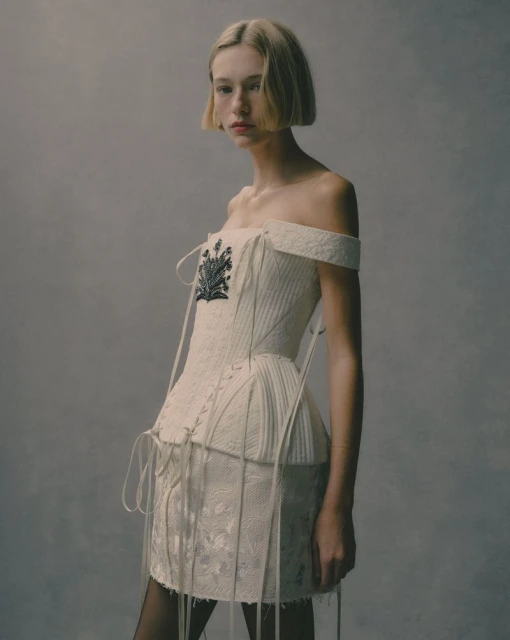Fashion shot of a young woman with blonde bob wearing a white, off-the-shoulder corset dress with black tights, posing in a studio.