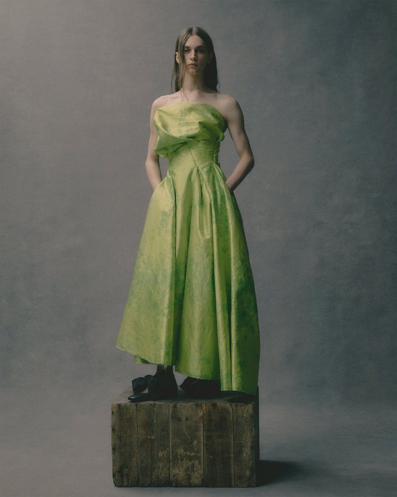 Model standing on a wooden crate wearing a strapless green dress against a muted textured background.