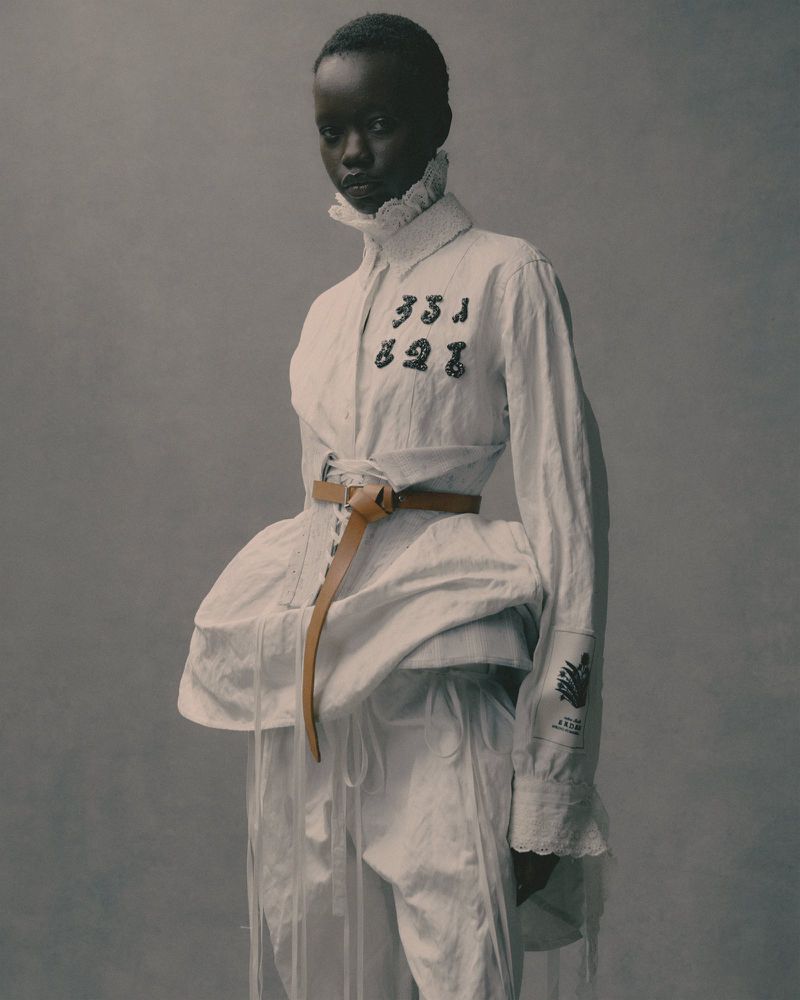 Fashionable African-descent woman in a white shirt with ruffle collar and tan belt, striking a pose against a neutral gray background.