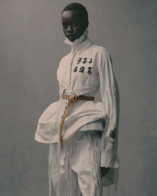 Fashionable African-descent woman in a white shirt with ruffle collar and tan belt, striking a pose against a neutral gray background.