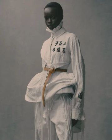 Fashionable African-descent woman in a white shirt with ruffle collar and tan belt, striking a pose against a neutral gray background.