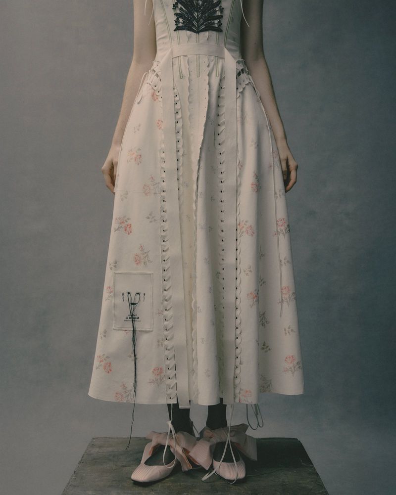 Person wearing floral dress and pink ballet shoes standing on a wooden platform in a photo studio setting, fashion editorial.