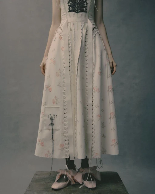 Person wearing floral dress and pink ballet shoes standing on a wooden platform in a photo studio setting, fashion editorial.