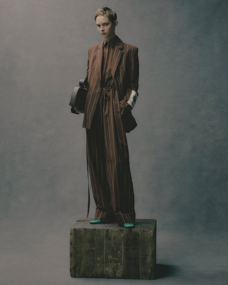 An androgynous model poses on a wooden block wearing a brown pinstripe suit and holding a helmet in a grey photo studio setting.