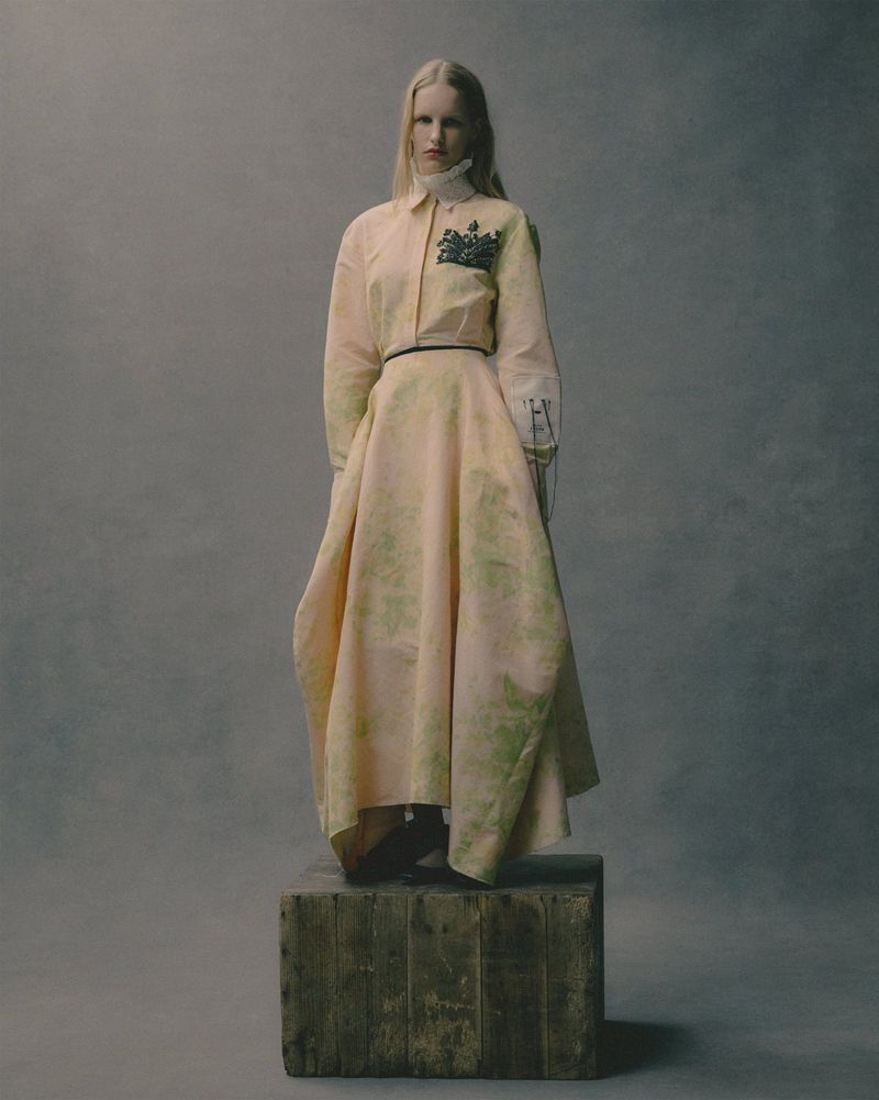 A woman in a long, patterned dress stands on a wooden crate in a studio setting. The dress features pale hues and a dark belt.