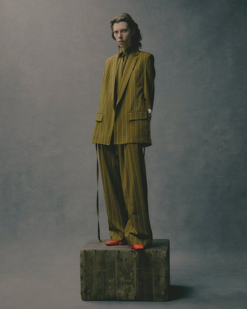 A slender figure stands on a wooden cube, sporting a striped olive-green suit with matching shirt and vibrant orange shoes in a studio shot.