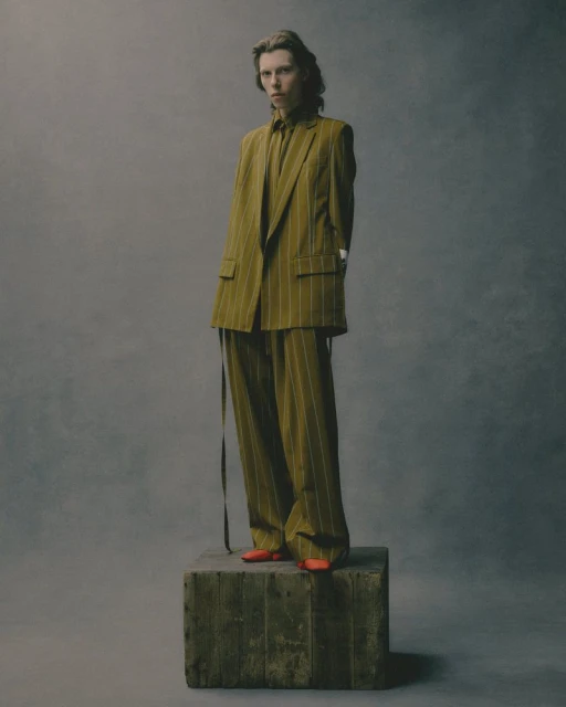A slender figure stands on a wooden cube, sporting a striped olive-green suit with matching shirt and vibrant orange shoes in a studio shot.