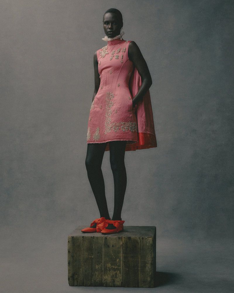 Elegant African-American woman in a stylish pink dress, black tights, and red shoes, standing gracefully against a grey backdrop.